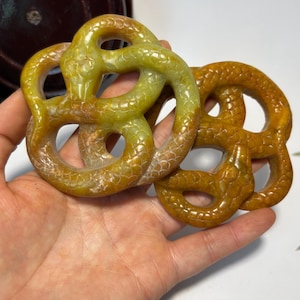 May include: Two circular carved stone pendants featuring an intertwined snake design. The left pendant is a mottled light green and yellow, while the right pendant is a solid deep amber-yellow. Both pieces display detailed scale textures.