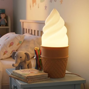 May include: A table lamp shaped like a soft serve ice cream cone. The top portion is a white, glowing, swirled shade, while the base is a tan, textured waffle cone. The lamp emits a warm, soft light and includes a power cord extending from the side.