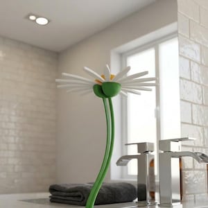 Puede incluir: Un accesorio de baño decorativo con forma de margarita con pétalos blancos, centro amarillo y tallo verde. La margarita está colocada frente a un lavabo con grifos cromados y un dispensador de jabón marrón.