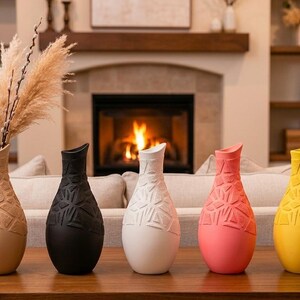 May include: Five decorative vases in a row, each with a unique color and geometric design. Colors include tan, black, white, coral, and yellow. The tan vase holds dried ornamental grass. The vases are on a wooden table.