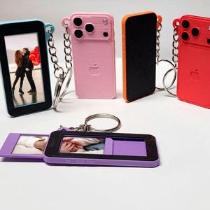 May include: A selection of colourful keychain phone cases, including pink, red, and blue, with a photo display. Each case has a silver chain and a keyring. The cases resemble a smartphone design.