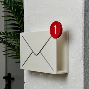 Puede incluir: Un organizador de correo de pared blanco con forma de sobre y una insignia de notificación roja con el número 1 en blanco. El organizador incluye tres pequeños ganchos en la parte inferior para colgar objetos y una ranura de almacenamiento.
