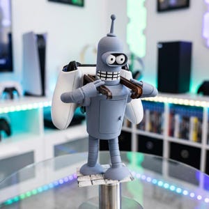 May include: A gray robot figurine with a white controller on each side. The robot has a toothy grin and is holding a brown tool. The figurine is standing on a small platform.