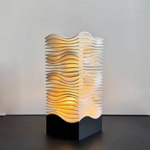 Puede incluir: Una lámpara de mesa con una base rectangular negra y una estructura vertical compuesta por capas horizontales blancas, onduladas y apiladas. El interior de la lámpara emite una luz amarilla cálida y suave que ilumina los espacios entre las capas.