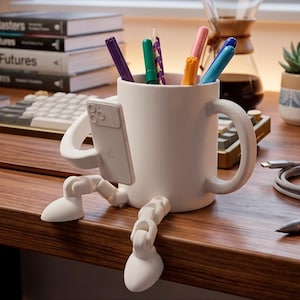 May include: A white ceramic mug with a unique design, holding various colored pens and pencils. The mug has arms and legs, and is holding a phone. The mug is on a wooden desk, with books and a keyboard in the background.