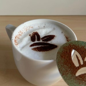 May include: A white ceramic mug filled with a cappuccino, featuring a cocoa powder design of a face. A green and brown stencil with a similar face design is next to the mug. The coffee art is on a light wooden surface.