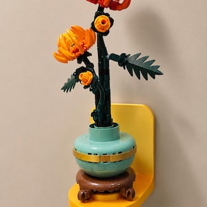 Könnte beinhalten: Ein aus Kunststoffbausteinen gefertigtes Blumenarrangement mit drei orange-gelben, chrysanthemenartigen Blüten an dunkelgrünen Stielen mit gezackten Blättern, in einer hellblauen Schale auf einem braunen runden Sockel, an einem gelben Wandregal.