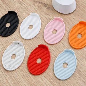 Può includere: Un assortimento di saponette ovali in silicone nei colori nero, bianco, rosa, arancione, bianco maculato, rosso e azzurro. Ogni saponetta ha un foro centrale e un bordo rialzato. Disposte su una superficie in legno chiaro.