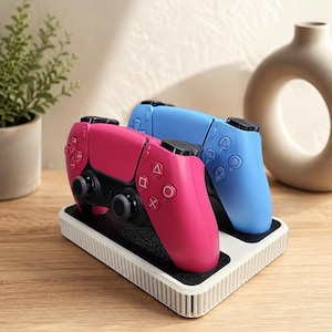 May include: A white rectangular charging dock with a black interior base holding two wireless game controllers, one in bright pink and one in light blue, each featuring black analog sticks and standard geometric action buttons on the front.