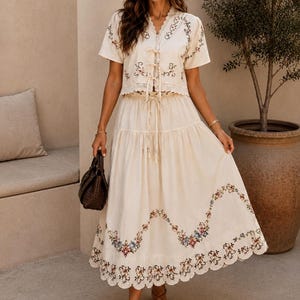 May include: A cream-colored two-piece set consisting of a short-sleeved blouse with a V-neckline, front tie closures, and floral embroidery, paired with a matching tiered midi skirt featuring a scalloped hem and colorful floral embroidery.