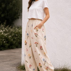 May include: A white short-sleeved crop top paired with beige high-waisted trousers featuring an elastic waistband, side pockets, and a scattered floral embroidery pattern in shades of blue, yellow, pink, and green, finished with cuffed ankles.