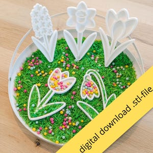 Op de afbeelding: Witte bloemvormige koekjessnijders op een bedje van groene hagelslag en kleurrijke snoepjes. De snijders omvatten een hyacint, narcis en krokus. De afbeelding heeft een gele diagonale banner met de tekst "digital download .stl-file".