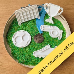 May include: A collection of white plastic sports-themed cookie cutters including a soccer goal, a trophy, a whistle, and a soccer cleat, alongside a bronze-colored soccer medal with a blue ribbon, all resting on a bed of green shredded material.
