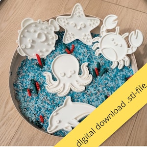 Op de afbeelding: Een set van vijf witte uitsteekvormpjes in de vorm van zeedieren, waaronder een kogelvis, zeester, krab, octopus en dolfijn, gerangschikt in een ronde schaal gevuld met blauwe en grijze rijst en rode pastastukjes. Tekst: digital download .stl-file.