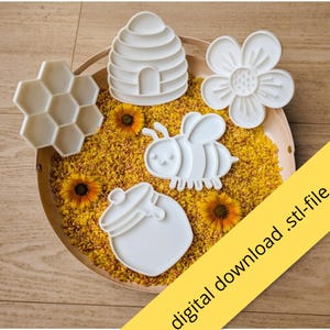 Op de afbeelding: Een set van vijf witte plastic koekjesvormen met een bijenkorf, bloem, bij, honingpot en honingraatpatroon. De items liggen op een ronde schaal gevuld met gele korrels en kleine bloemen. De tekst luidt: digital download .stl-file.