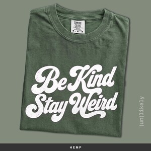 May include: A dark olive green crew neck t-shirt with white screen-printed text that reads Be Kind Stay Weird in a cursive font. The shirt features a Comfort Colors brand label at the inner neckline indicating a size medium and 100% cotton material.