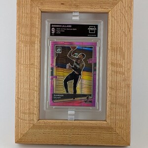 May include: A basketball trading card of Damian Lillard, graded as a 9, in a protective case, set within a light brown wooden frame. The card has a pink and gold design. Text includes "DAMIAN LILLARD" and "PORTLAND".