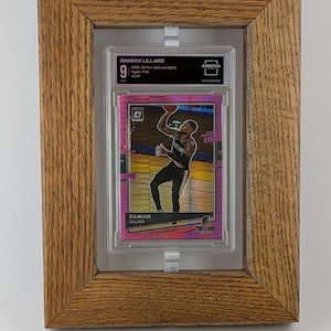 May include: A framed basketball card featuring Damian Lillard. The card is in a clear case, set against a pink backdrop. The frame is made of light brown wood. The text "Frame only. Card not included." is at the bottom of the image.