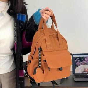 Könnte beinhalten: Ein orangefarbener Rucksack mit einer Vordertasche und einem kleinen, befestigten Beutel. Der Rucksack hat zwei obere Griffe und verstellbare Schultergurte. Ein schwarz-weißes Seildetail akzentuiert die Seite. Der Rucksack besteht aus einem strapazierfähigen, wasserabweisenden Material.