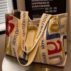 May include: Cream tote bag with colourful lettering and cream straps. The bag features the word "GENTLEWOMAN" in various colours. The straps also have the word "GENTLEWOMAN" printed on them. The bag is on a metallic surface.