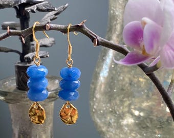 Könnte beinhalten: Blaue Perlenohrringe mit goldfarbenen Akzenten. Die Ohrringe hängen an einem Ast, jeweils mit einer goldfarbenen, gehämmerten Scheibe. Im Hintergrund befinden sich eine dekorative Vase und eine rosa Orchidee.