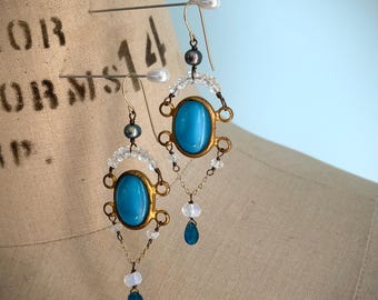 Könnte beinhalten: Ein Paar verzierte Ohrringe mit ovalen türkisfarbenen Steinen, die in goldfarbenem Metall gefasst sind. Die Ohrringe haben ein filigranes Design mit klaren und dunkelgrauen Perlen sowie tropfenförmigen blauen Akzenten. Die Ohrringe werden auf einer Schaufensterpuppe präsentiert.