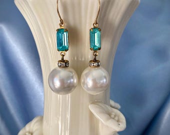 Könnte beinhalten: Ein Paar elegante Ohrringe mit goldfarbenen Haken. Jeder Ohrring hat einen rechteckigen, hellblauen Edelstein über einer großen, weißen Perle. Die Edelsteine sind mit einem kleinen, dekorativen Element mit den Perlen verbunden.