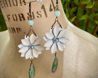 Könnte beinhalten: Weiße Blumenohrringe mit blauem Mittelstein und tropfenförmigem grün-violettem Stein. Die Ohrringe haben eine Kette und Perlendetails und werden auf einer Schaufensterpuppe präsentiert.