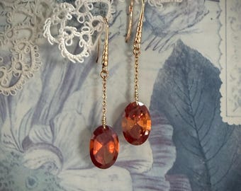 Könnte beinhalten: Goldfarbene Ohrhänger mit ovalen, orange-roten Edelsteinen. Die Ohrringe hängen an feinen Ketten. Der Hintergrund zeigt ein Blumenmuster und eine zarte Spitze.