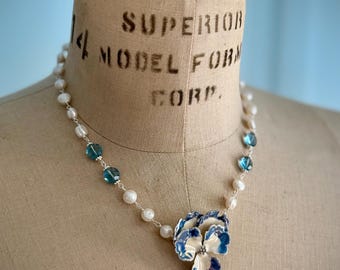 Könnte beinhalten: Zarte Halskette mit einem floralen Anhänger mit blau-weißen Emaille-Details. Die Halskette enthält Süßwasserperlen und blaue Kristallperlen. Der Schmuck wird auf einer Vintage-Schneiderpuppe präsentiert.