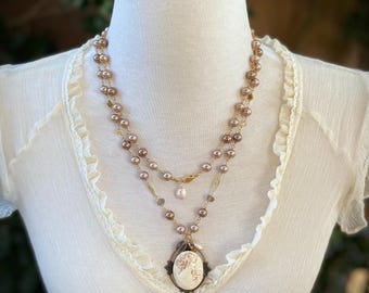 Könnte beinhalten: Eine mehrreihige Halskette mit einem Kameenanhänger. Die Halskette besteht aus braunen und goldenen Perlen und hat einen zarten Perlenakzent. Der Kameenanhänger ist ein weißes Porzellan-Design eines Frauenkopfes.