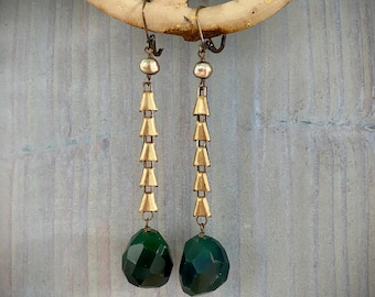Könnte beinhalten: Ein Paar lange Ohrhänger mit einem messingfarbenen Metallhaken und einer vertikalen Kette aus vier kleinen, dreieckigen, goldfarbenen Metallperlen. Jeder Ohrring endet in einer großen, facettierten, dunkelgrünen Edelsteinperle.