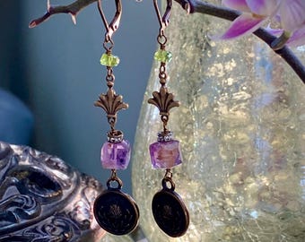 Könnte beinhalten: Hängeohrringe im Vintage-Stil. Jeder Ohrring hat einen kupferfarbenen Haken, grüne Perlen, verziertes Filigran, facettierte lila Edelsteine und einen dunklen runden Anhänger. Die Ohrringe hängen an einem Ast vor einem verschwommenen Hintergrund.