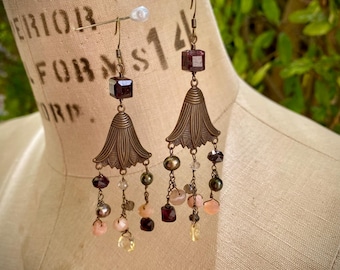 Könnte beinhalten: Ohrringe zum Hängen im Vintage-Stil. Die Ohrringe haben eine bronzefarbene, stilisierte Glockenform mit hängenden Perlen in Rosa, Lila und Grün. Jeder Ohrring hat einen quadratischen, dunkelvioletten Edelstein an der Spitze.