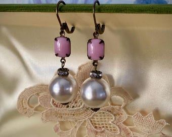 Könnte beinhalten: Ein Paar Ohrringe im Vintage-Stil. Jeder Ohrring hat einen hellrosa, quadratischen Stein, eine große weiße Perle und einen kleinen, dekorativen Metallakzent. Die Ohrringe hängen an einem gebogenen, antik aussehenden Metallhaken.
