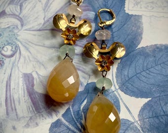 Könnte beinhalten: Ein Paar goldfarbene Ohrhänger mit floralem Design. Die Ohrringe haben facettierte, tropfenförmige gelbe Edelsteine, kleine klare Perlen und goldene Blumenakzente. Der Hintergrund ist ein blau-weißes Blumenmuster.