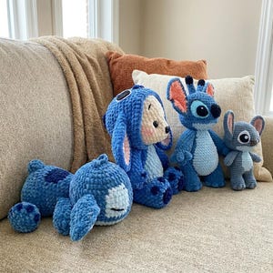 K&ouml;nnte beinhalten: Eine Sammlung von drei handgefertigten H&auml;kel-Pl&uuml;schfiguren, die den Charakter Stitch darstellen. Eine Figur liegt, eine sitzt mit blauer Kapuze und eine steht. Die Figuren sind aus weichem blauem Garn mit rosafarbenen Innenohren gefertigt.