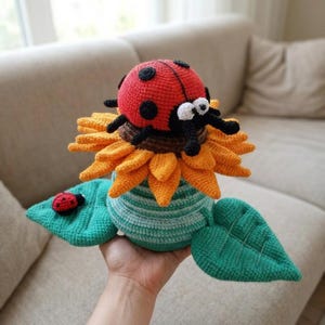 May include: A crocheted stuffed toy featuring a bright red ladybug with black spots resting on a large orange sunflower. The flower sits atop a teal and white striped base with two large green leaves, one of which holds a tiny red ladybug.