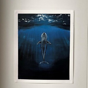 May include: An original painting of a whale swimming in deep blue water. The whale is depicted from above, with a white and grey body. The water is dark blue, with light rays shining down from the surface. The artwork is on white paper.