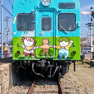 以下が含まれることがあります： 前面に漫画のキャラクターが描かれた、ターコイズとグリーンの電車。 列車は砂利のベッドのある線路にあります。 空は青く、雲がいくつかあります。