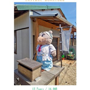 以下が含まれることがあります： 白いシャツ、赤いネクタイ、水色のズボンを着用し、マスクと頭巾を被ったクマのぬいぐるみ。木製の台の上に立ち、横には木箱と縦型の旗が置かれている。テキストには「LITTLE SANCTUARY: THE BEAR GUARDIAN」と書かれている。