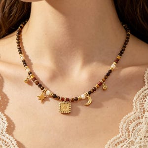Op de afbeelding: Een kralenketting met een hemels thema. De ketting heeft een mix van bruine, witte en gouden kralen. Gouden bedels zijn onder meer een ster, een halve maan en een vierkante hanger met een zonmotief. Kleine gouden balbedels zijn ook inbegrepen.