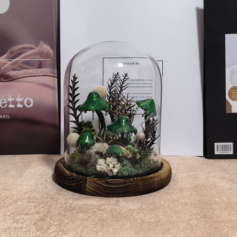 Pu&ograve; includere: Una campana di vetro con base in legno ospita una scena forestale in miniatura. La scena comprende figurine di funghi verdi, piante secche e fiori bianchi. La campana poggia su una base rotonda in legno marrone scuro. La parola "DREAMING" &egrave; visibile.