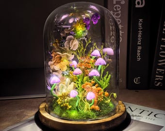 May include: A glass dome cloche on a circular wooden base containing a miniature forest scene with preserved flowers, green moss, and several small, glowing purple mushroom-shaped lights that illuminate the arrangement from within the display.