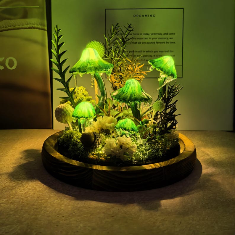 Pu&ograve; includere: Un terrario decorativo di funghi illuminati. I funghi di vetro brillano di verde, incastonati tra muschio, piante e fiori in una ciotola di legno. Lo sfondo include un foglio con la parola "DREAMING".