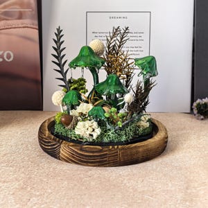 Pu&ograve; includere: Un terrario decorativo con funghi di vetro verde, fiori bianchi e fogliame secco, posto all'interno di una base rotonda in legno. La composizione comprende varie trame e colori, creando una scena di foresta in miniatura. Lo sfondo include una stampa incorniciata con la parola "DREAMING".