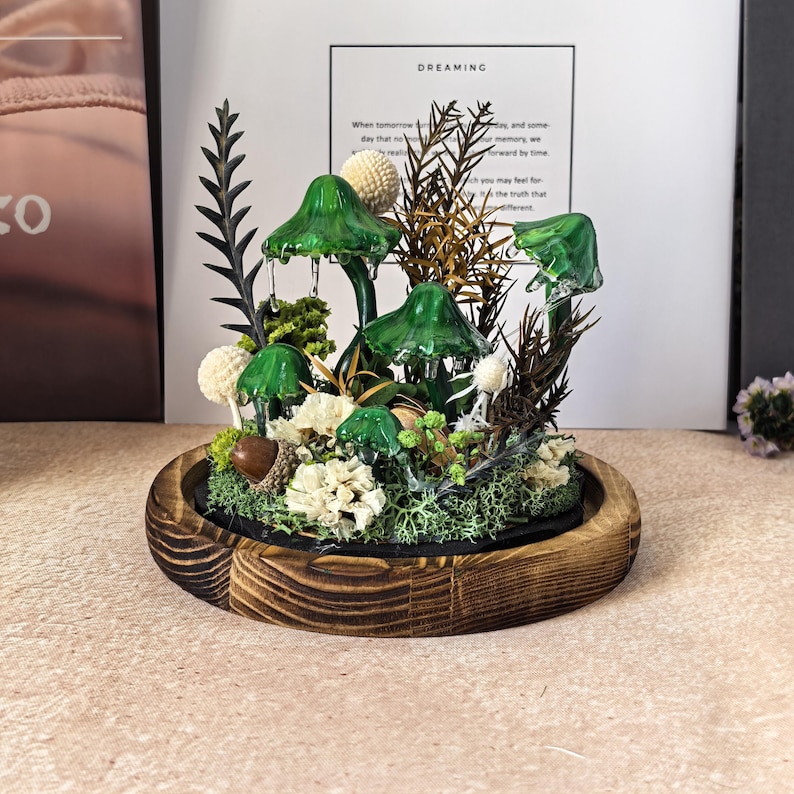 Pu&ograve; includere: Un terrario decorativo con funghi di vetro verde, fiori bianchi e fogliame secco, posto all'interno di una base rotonda in legno. La composizione comprende varie trame e colori, creando una scena di foresta in miniatura. Lo sfondo include una stampa incorniciata con la parola "DREAMING".