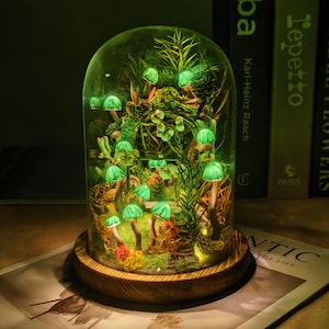 Puede incluir: Un terrario de cúpula de cristal sobre una base circular de madera que contiene una escena de bosque en miniatura con múltiples luces verdes brillantes en forma de seta, varios musgos verdes, ramitas y follaje, creando un decorado boscoso.