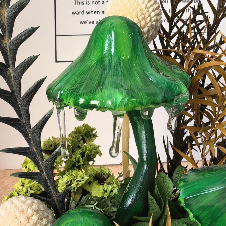 Pu&ograve; includere: Una scultura a forma di fungo verde realizzata a mano con un cappello strutturato e accenti trasparenti gocciolanti. Il fungo &egrave; circondato da fogliame secco e verde, creando un'esposizione naturalistica. Il testo sullo sfondo &egrave; parzialmente visibile.