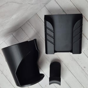 Puede incluir: Un conjunto de tres objetos negros impresos en 3D. El conjunto incluye un soporte en forma de taza, un soporte rectangular con líneas anguladas y una pieza pequeña y curvada. Los objetos están dispuestos sobre una superficie clara con vetas de madera.